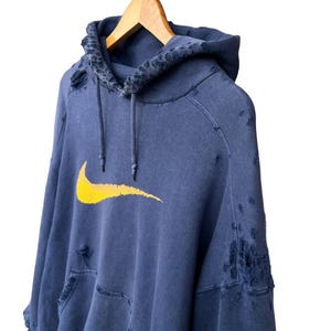 Puede incluir: Una sudadera azul desgastada con un logotipo de Nike swoosh amarillo. La sudadera tiene varios agujeros y desgarros en toda la tela.