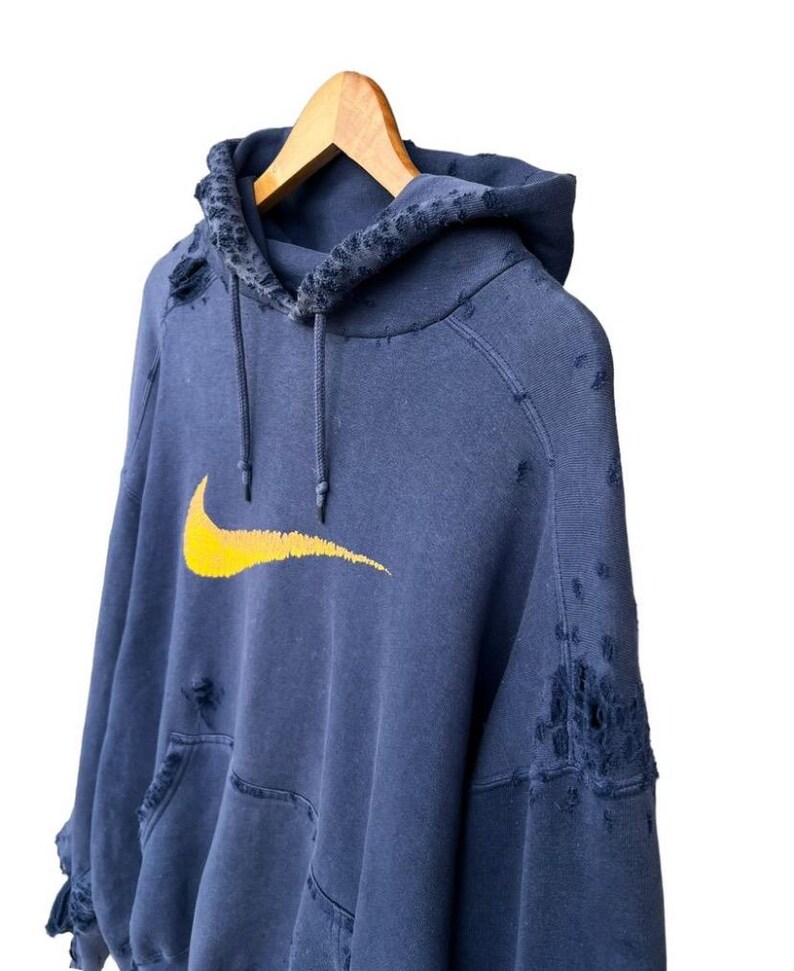 Puede incluir: Una sudadera azul desgastada con un logotipo de Nike swoosh amarillo. La sudadera tiene varios agujeros y desgarros en toda la tela.
