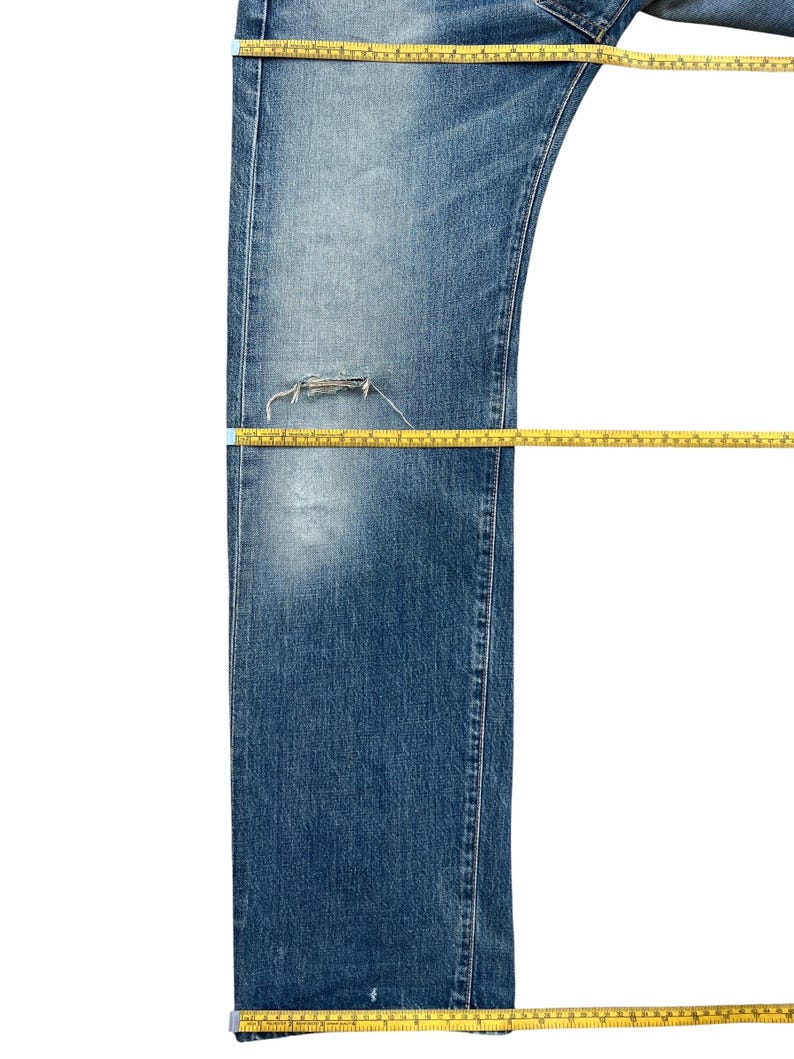Puede incluir: Vaqueros azules de mezclilla con una zona de rodilla desgastada. Una cinta m&eacute;trica amarilla se coloca sobre los vaqueros para medir la longitud. Los vaqueros tienen un lavado descolorido y un dise&ntilde;o cl&aacute;sico de cinco bolsillos.