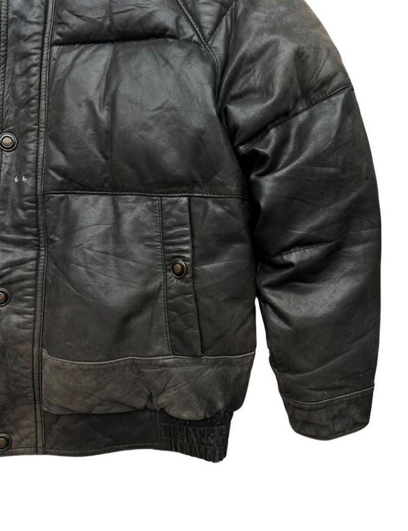 May include: Close-up of a black leather jacket with a quilted design. The jacket features a button-up front, a pocket on the chest, and a ribbed hem. The leather has a worn, distressed appearance.