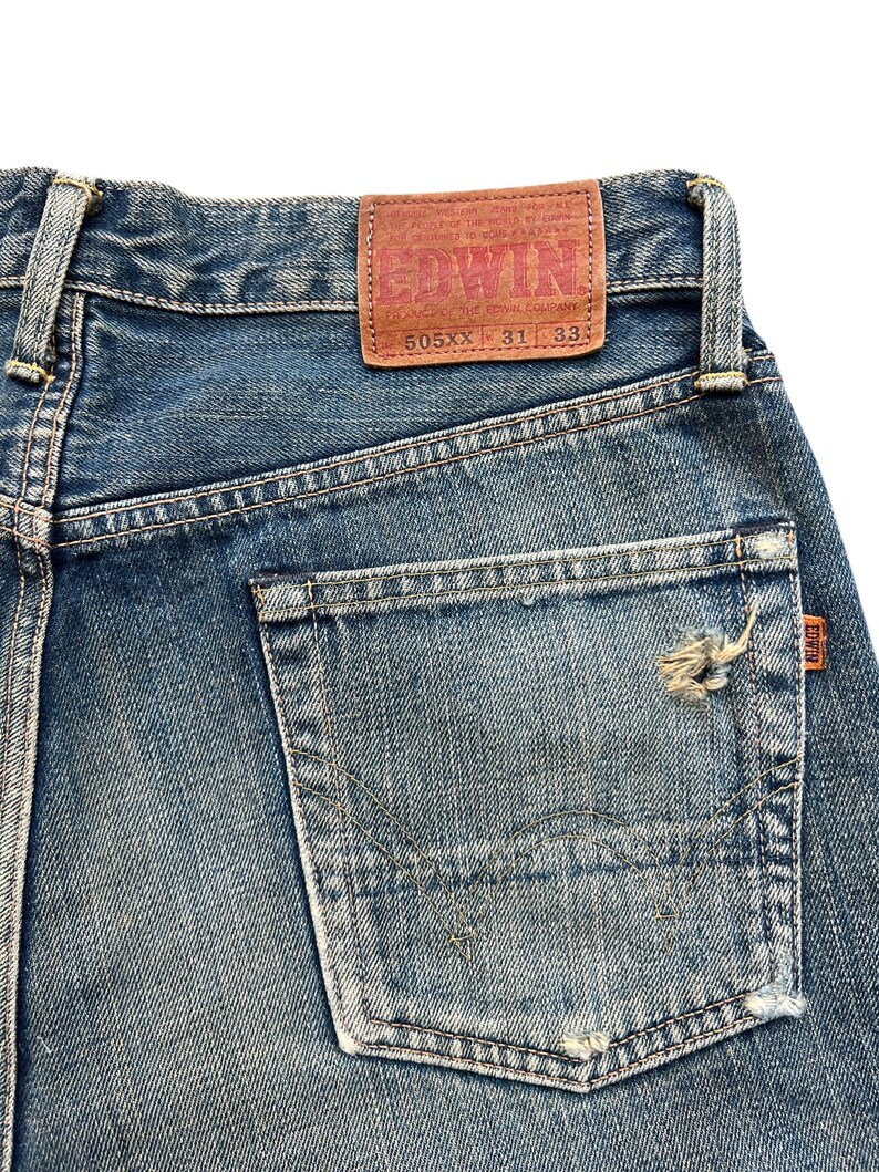 May include: Close-up of blue denim jeans with a distressed pocket. A brown leather patch with the brand name "EDWIN" is visible. The jeans are labeled "505XX 31 33".