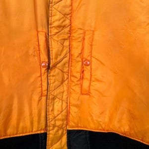 May include: Close-up of a bright orange bomber jacket. The jacket features two front pockets with orange buttons, a black ribbed hem, and a black zipper. The jacket has a quilted design along the zipper and pocket seams.