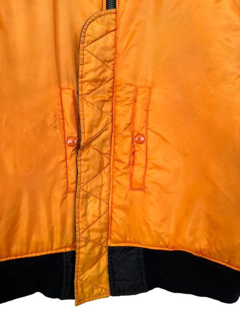 May include: Close-up of a bright orange bomber jacket. The jacket features two front pockets with orange buttons, a black ribbed hem, and a black zipper. The jacket has a quilted design along the zipper and pocket seams.
