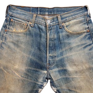 May include: Close-up of vintage blue denim jeans with a button fly. The jeans show signs of wear, with faded areas and distressed details. Features include classic five-pocket styling and copper rivets.