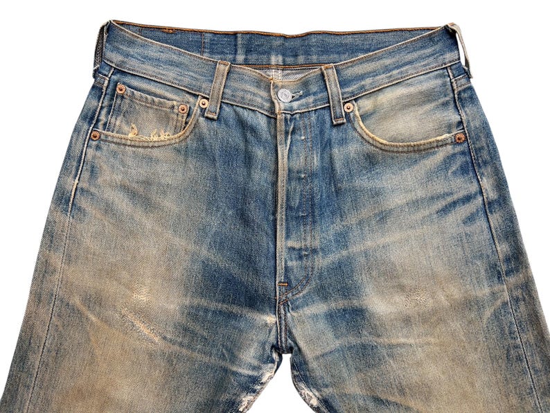 May include: Close-up of vintage blue denim jeans with a button fly. The jeans show signs of wear, with faded areas and distressed details. Features include classic five-pocket styling and copper rivets.