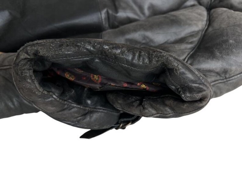 May include: Close-up of a black leather jacket sleeve. The leather shows signs of wear and tear, with a distressed appearance. The interior lining is visible, featuring a patterned design. A buckle detail is present near the cuff.
