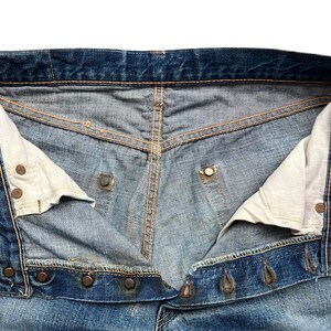 以下が含まれることがあります： 青いデニムジーンズの開いたウエストバンドのクローズアップ。ジーンズは金属製のボタンが付いたボタンフライで、内側の裏地が見えます。デニムは使い古された、色あせた外観をしています。