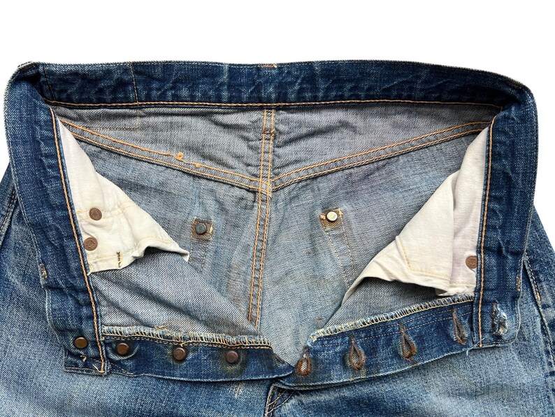 以下が含まれることがあります： 青いデニムジーンズの開いたウエストバンドのクローズアップ。ジーンズは金属製のボタンが付いたボタンフライで、内側の裏地が見えます。デニムは使い古された、色あせた外観をしています。