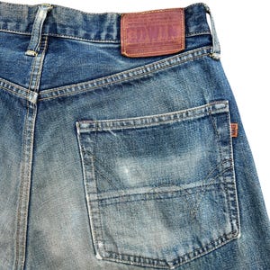May include: Close-up of the back of blue denim jeans. The jeans have a faded wash and a large back pocket. A brown leather patch with the brand name "EDWIN" is sewn on the waistband.
