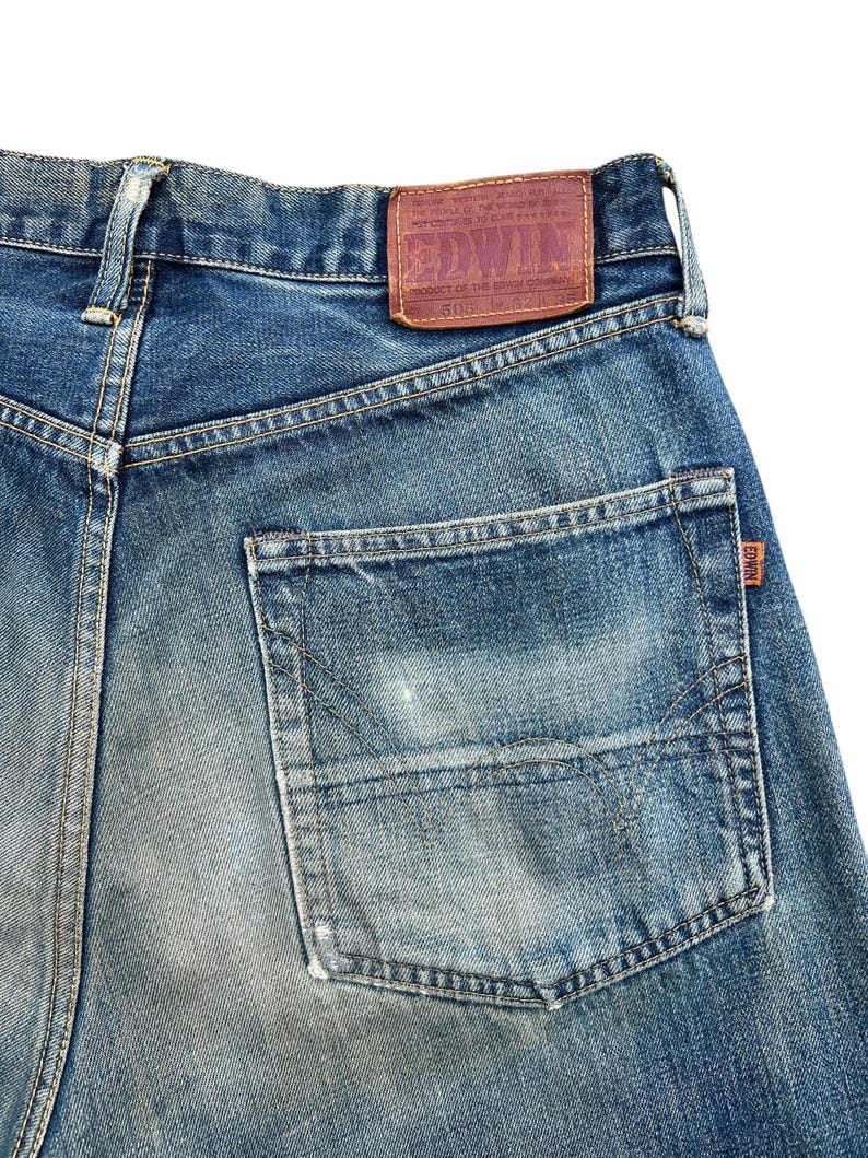 May include: Close-up of the back of blue denim jeans. The jeans have a faded wash and a large back pocket. A brown leather patch with the brand name "EDWIN" is sewn on the waistband.