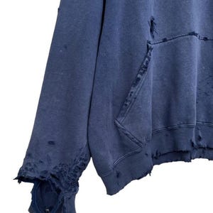 Puede incluir: Una sudadera azul desgastada con un estampado gr&aacute;fico amarillo. La sudadera tiene varios agujeros y desgarros en toda la tela.