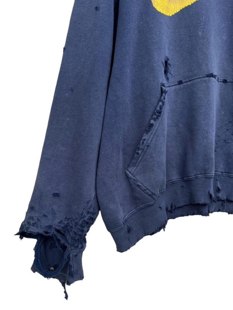 Puede incluir: Una sudadera azul desgastada con un estampado gr&aacute;fico amarillo. La sudadera tiene varios agujeros y desgarros en toda la tela.