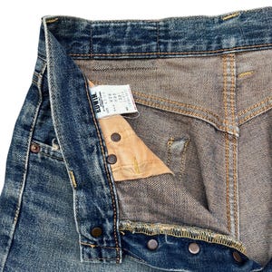 Puede incluir: Primer plano de unos vaqueros de mezclilla azules con bragueta de botones. Los vaqueros tienen un aspecto desgastado y descolorido con costuras y remaches met&aacute;licos visibles. Se ve una etiqueta con la marca "Edwin".