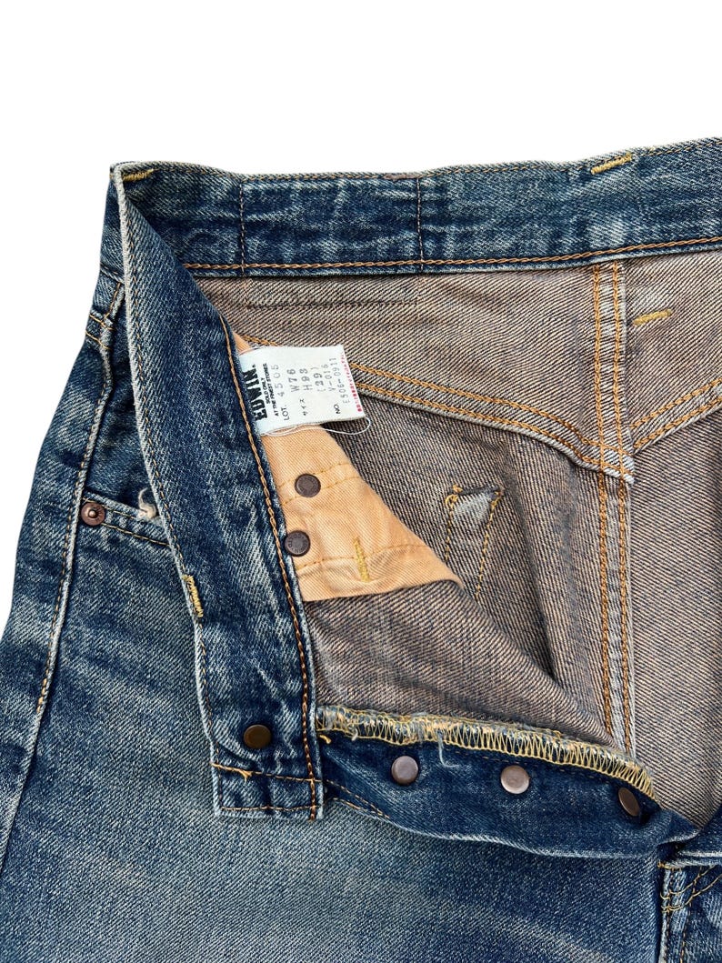 Puede incluir: Primer plano de unos vaqueros de mezclilla azules con bragueta de botones. Los vaqueros tienen un aspecto desgastado y descolorido con costuras y remaches met&aacute;licos visibles. Se ve una etiqueta con la marca "Edwin".