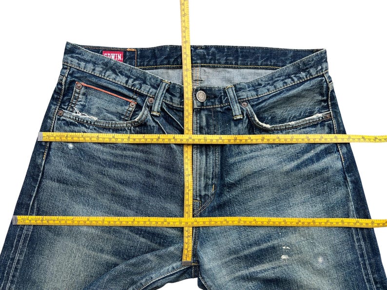 Puede incluir: Primer plano de unos vaqueros de mezclilla azules con un lavado descolorido y detalles desgastados. Los vaqueros tienen un dise&ntilde;o cl&aacute;sico de cinco bolsillos, cierre de bot&oacute;n y una etiqueta roja. Una cinta m&eacute;trica amarilla est&aacute; colocada sobre los vaqueros.
