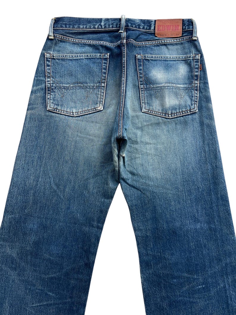 Puede incluir: Vaqueros de mezclilla azul descolorido con dos bolsillos traseros y un parche de cuero. Los vaqueros tienen un dise&ntilde;o cl&aacute;sico de cinco bolsillos y un corte recto. El nombre de la marca "EDWIN" es visible en el parche de cuero.