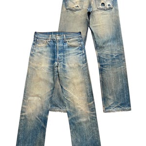 May include: Two pairs of vintage blue denim jeans. The jeans have a faded wash, distressed details, and visible wear and tear, including holes and frayed edges. One pair is laid flat, while the other is positioned behind it.