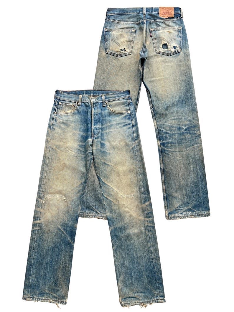 May include: Two pairs of vintage blue denim jeans. The jeans have a faded wash, distressed details, and visible wear and tear, including holes and frayed edges. One pair is laid flat, while the other is positioned behind it.