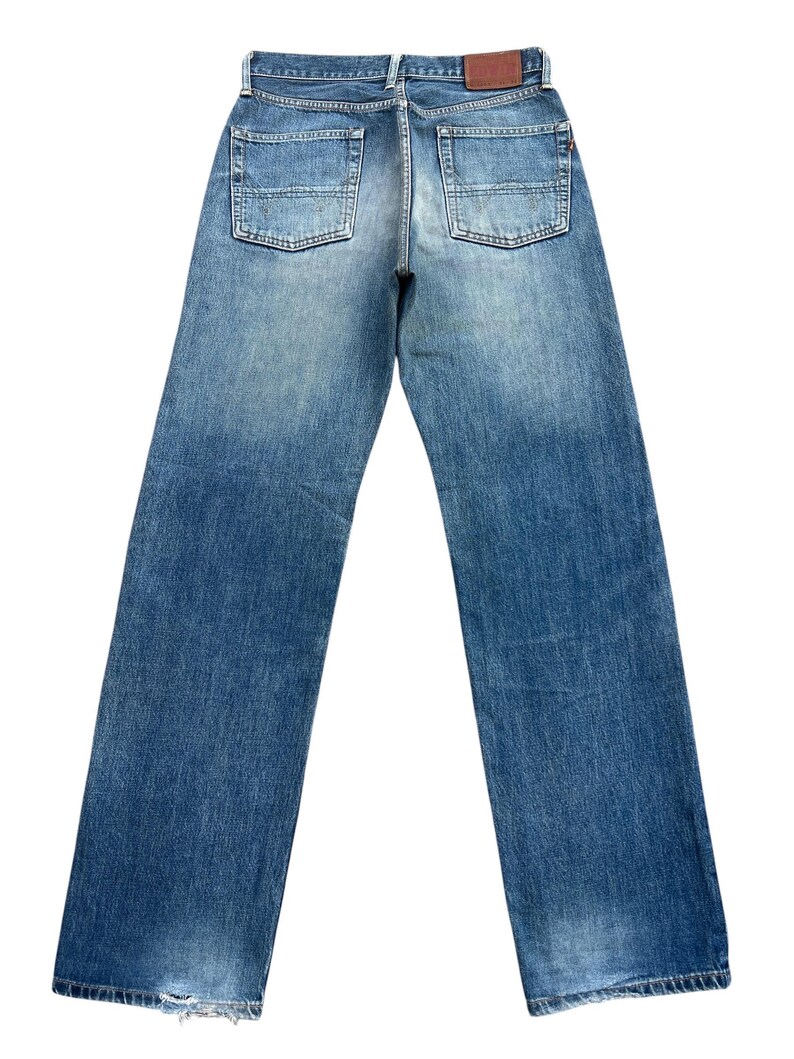 Puede incluir: Vista trasera de unos vaqueros de mezclilla azules con dos bolsillos traseros y un parche de cuero marr&oacute;n. Los vaqueros tienen un lavado descolorido y un dise&ntilde;o de pernera recta. El dobladillo inferior de los vaqueros est&aacute; ligeramente desgastado.