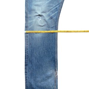 Pu&ograve; includere: Jeans di denim blu dall'aspetto usurato, con un'area usurata e uno strappo. I nastri di misurazione gialli sono posizionati sui jeans per la misurazione. I jeans sono su uno sfondo bianco.