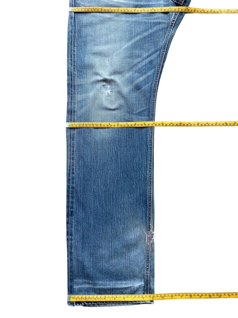 Pu&ograve; includere: Jeans di denim blu dall'aspetto usurato, con un'area usurata e uno strappo. I nastri di misurazione gialli sono posizionati sui jeans per la misurazione. I jeans sono su uno sfondo bianco.