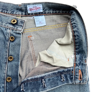 May include: Close-up of blue denim jeans with a button fly. The jeans have a faded wash and orange stitching. The label reads "Authentic Jeans Lady Wrangler Western Style". The waist size is 31 inches.