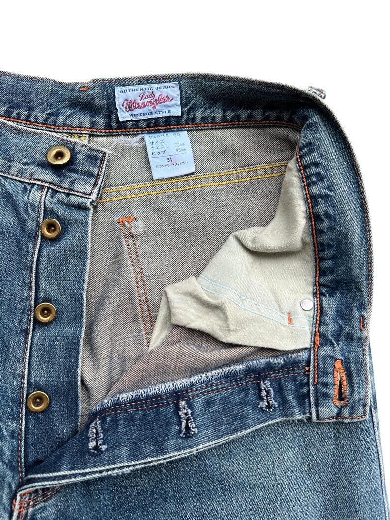 May include: Close-up of blue denim jeans with a button fly. The jeans have a faded wash and orange stitching. The label reads "Authentic Jeans Lady Wrangler Western Style". The waist size is 31 inches.
