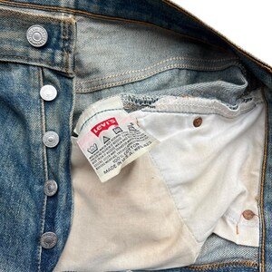 May include: Close-up of vintage blue denim jeans with a button fly. The Levi's brand label is visible inside the jeans. The label reads "100% Cotton" and "Made in U.S.A."