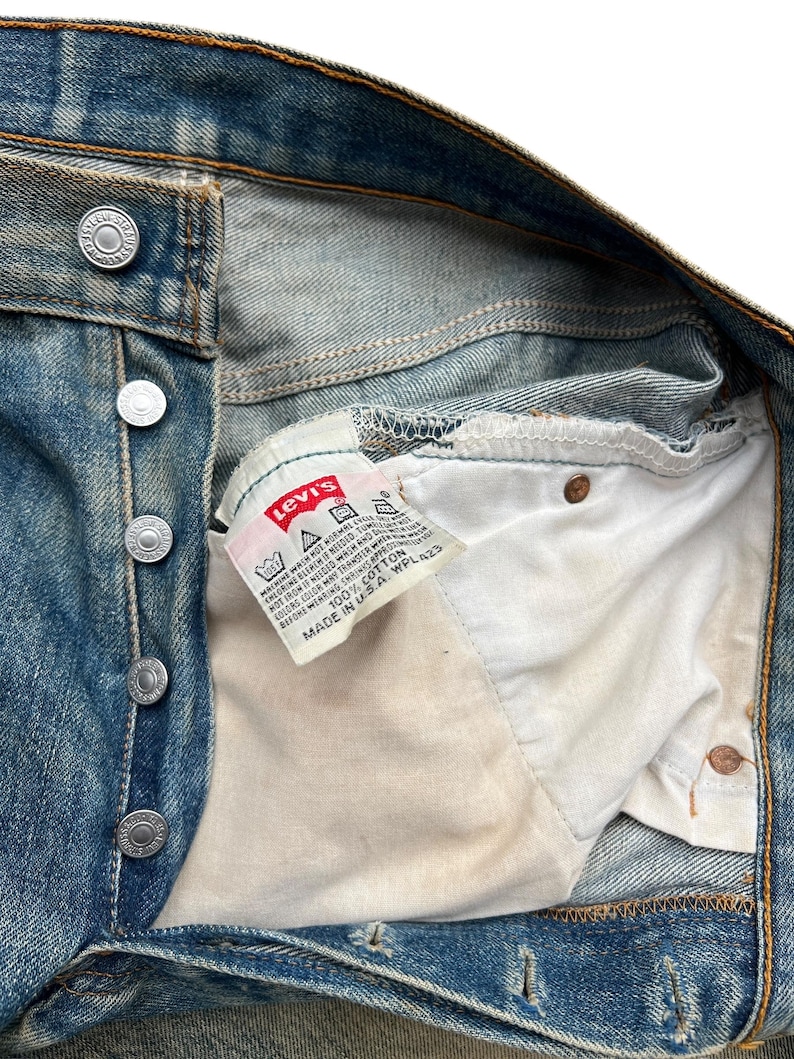 May include: Close-up of vintage blue denim jeans with a button fly. The Levi's brand label is visible inside the jeans. The label reads "100% Cotton" and "Made in U.S.A."