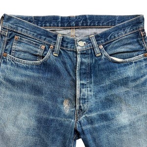 以下が含まれることがあります： ヴィンテージの青いデニムジーンズのクローズアップ。ジーンズはボタンフライ、クラシックな5ポケットデザインで、摩耗や色あせの兆候が見られます。デニムは、摩耗の跡や前面の明るい斑点が見られる、使い古された外観をしています。