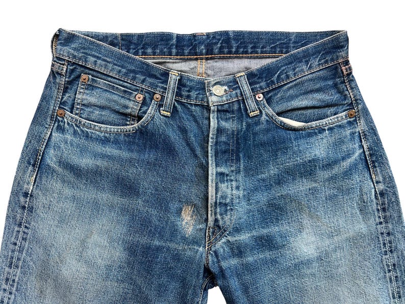 以下が含まれることがあります： ヴィンテージの青いデニムジーンズのクローズアップ。ジーンズはボタンフライ、クラシックな5ポケットデザインで、摩耗や色あせの兆候が見られます。デニムは、摩耗の跡や前面の明るい斑点が見られる、使い古された外観をしています。