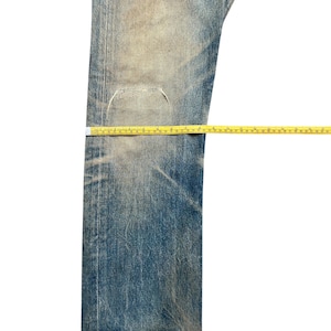 May include: Faded blue denim jeans with a distressed appearance. The jeans have a worn look with visible wear and tear, and are measured with a yellow measuring tape. The jeans are a classic style.