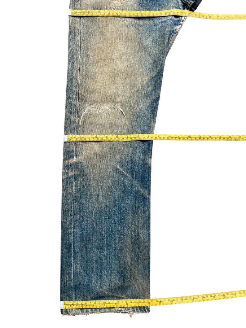 May include: Faded blue denim jeans with a distressed appearance. The jeans have a worn look with visible wear and tear, and are measured with a yellow measuring tape. The jeans are a classic style.