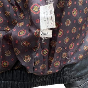 May include: Close-up of the inside of a black leather jacket. The lining is a dark brown fabric with a repeating pattern of red, gold, and black geometric shapes. A tag with Japanese text and the word "Duck" is visible.