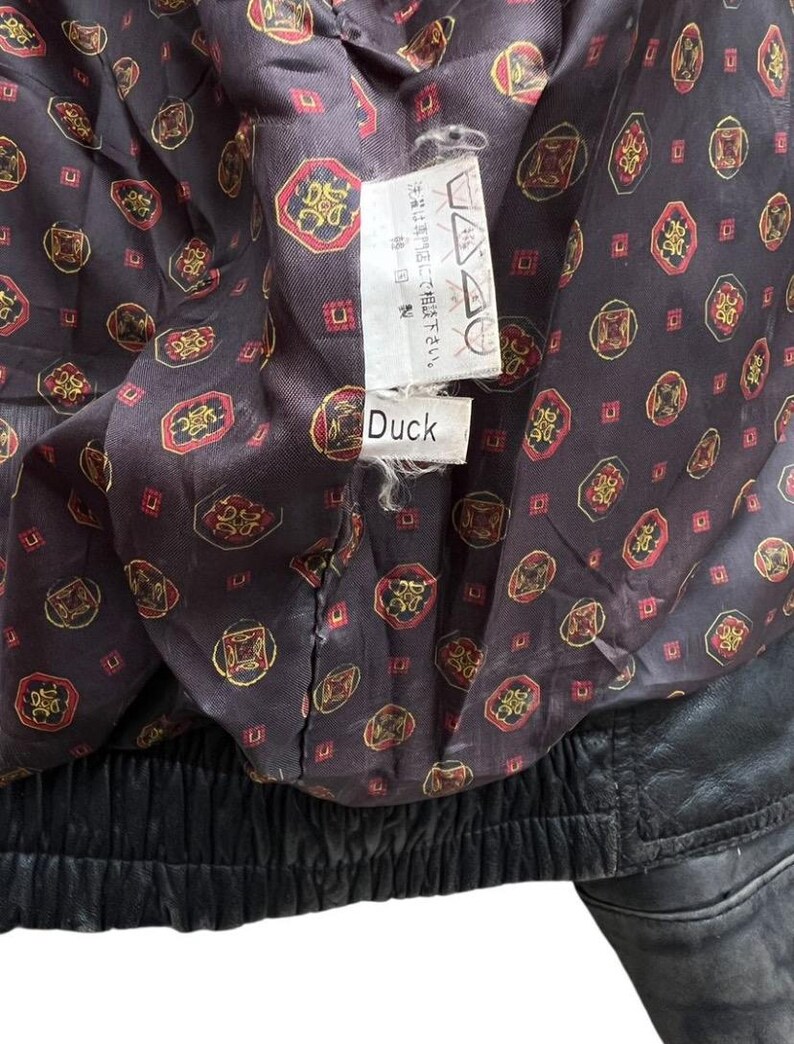 May include: Close-up of the inside of a black leather jacket. The lining is a dark brown fabric with a repeating pattern of red, gold, and black geometric shapes. A tag with Japanese text and the word "Duck" is visible.