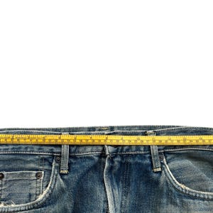 May include: Close-up of the waistband of blue denim jeans with a yellow measuring tape laid across the top. The jeans have a classic five-pocket design and show signs of wear.