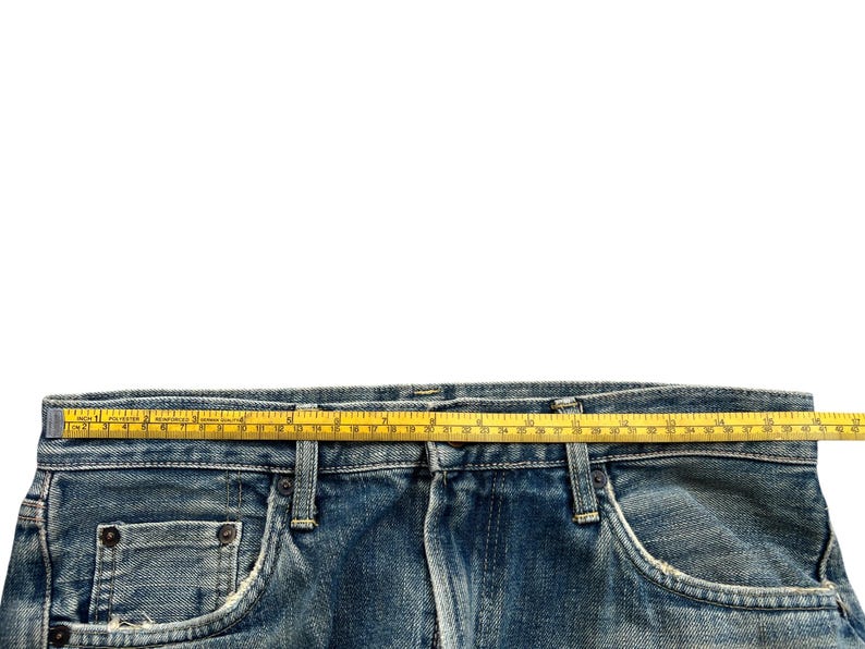 May include: Close-up of the waistband of blue denim jeans with a yellow measuring tape laid across the top. The jeans have a classic five-pocket design and show signs of wear.