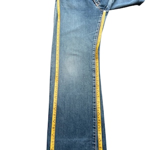 May include: Blue denim jeans with a straight leg design. A yellow measuring tape is placed along the side of the jeans to measure the length. The jeans are folded over on a white background.