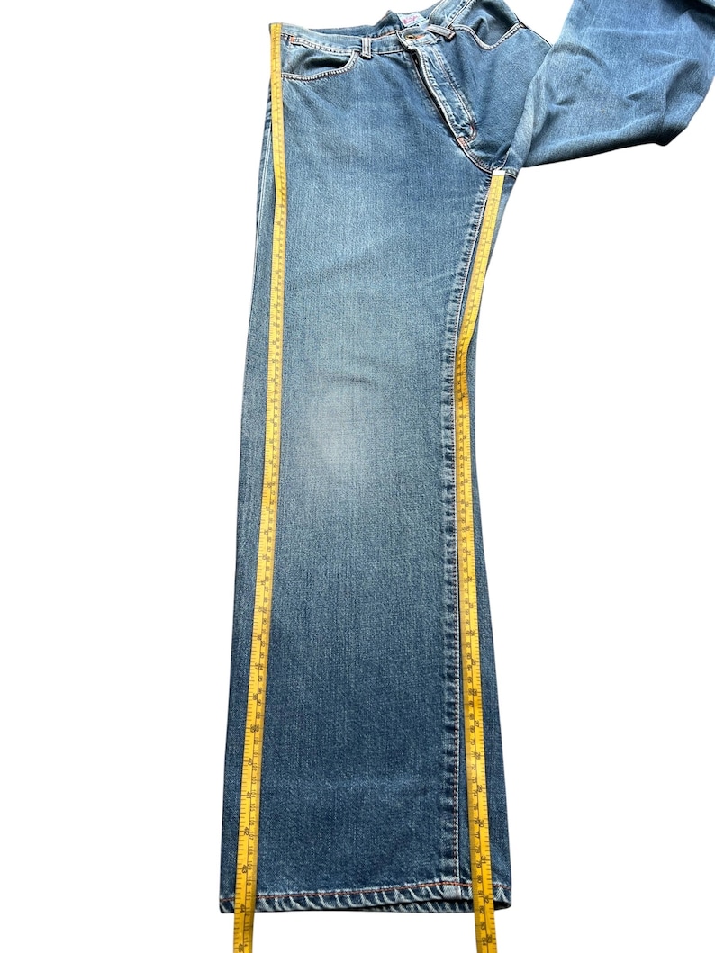 May include: Blue denim jeans with a straight leg design. A yellow measuring tape is placed along the side of the jeans to measure the length. The jeans are folded over on a white background.