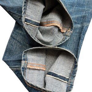 May include: Close-up of the folded cuffs of blue denim jeans. The cuffs are turned up to reveal the selvedge edge with a white and blue stripe and a contrasting orange stitch.