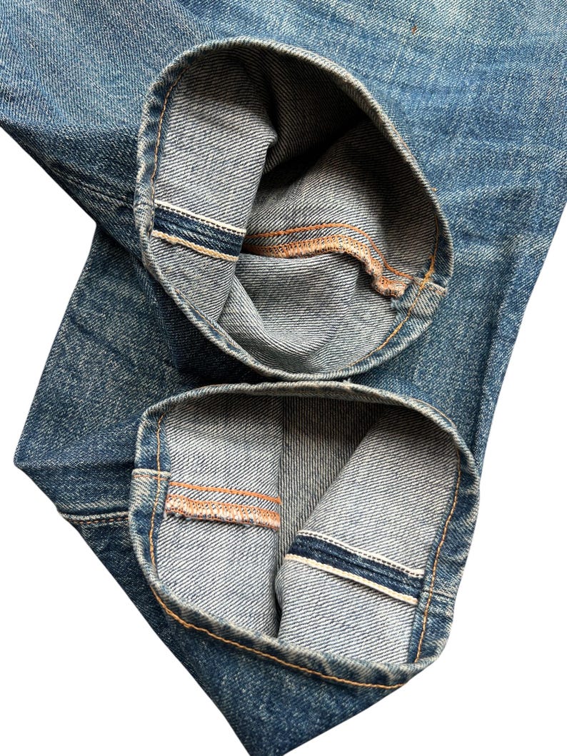 May include: Close-up of the folded cuffs of blue denim jeans. The cuffs are turned up to reveal the selvedge edge with a white and blue stripe and a contrasting orange stitch.