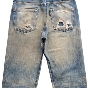 May include: Faded blue denim jeans with distressed details, including rips on the back pockets. The Levi Strauss & Co. label is visible on the back waistband. The jeans have a classic five-pocket design.