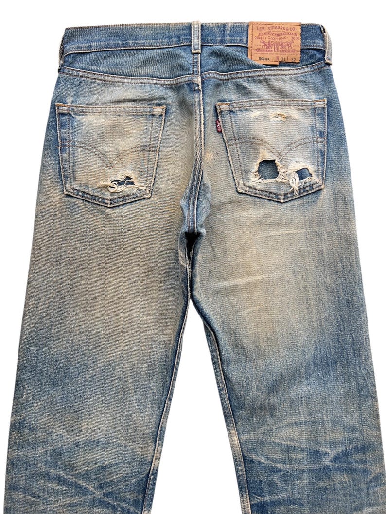 May include: Faded blue denim jeans with distressed details, including rips on the back pockets. The Levi Strauss & Co. label is visible on the back waistband. The jeans have a classic five-pocket design.