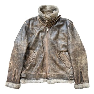 Könnte beinhalten: Eine braune Lederjacke im Used-Look mit Schafsfellkragen und -futter. Die Jacke hat einen Reißverschluss vorne, zwei Vordertaschen und Reißverschlussmanschetten. Die Jacke hat einen abgenutzten, Vintage-Look.