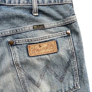 K&ouml;nnte beinhalten: Nahaufnahme der Ges&auml;&szlig;tasche einer blauen Wrangler-Jeans. Die Tasche hat einen braunen Lederaufn&auml;her mit dem Wrangler-Logo und der Aufschrift "AUTHENTIC WESTERN WEAR". Die Jeans hat ein klassisches F&uuml;nf-Taschen-Design.