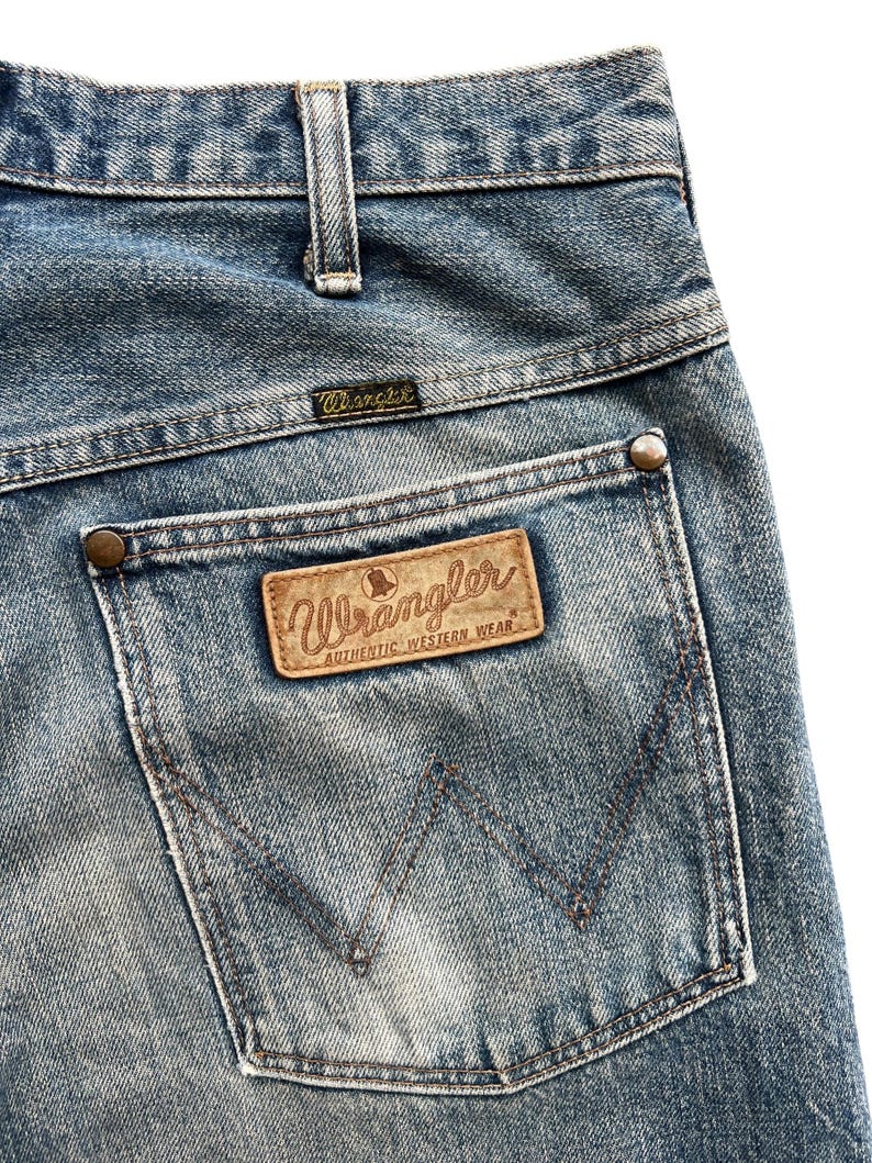 K&ouml;nnte beinhalten: Nahaufnahme der Ges&auml;&szlig;tasche einer blauen Wrangler-Jeans. Die Tasche hat einen braunen Lederaufn&auml;her mit dem Wrangler-Logo und der Aufschrift "AUTHENTIC WESTERN WEAR". Die Jeans hat ein klassisches F&uuml;nf-Taschen-Design.