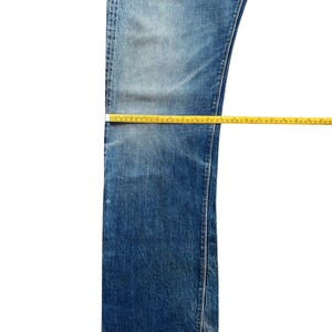 以下が含まれることがあります： 色あせたウォッシュ加工の青いデニムジーンズ。黄色のメジャーで測定されています。ジーンズは白い背景に平らに置かれ、片方の脚の前面が表示されています。メジャーはジーンズの長さと幅を示しています。