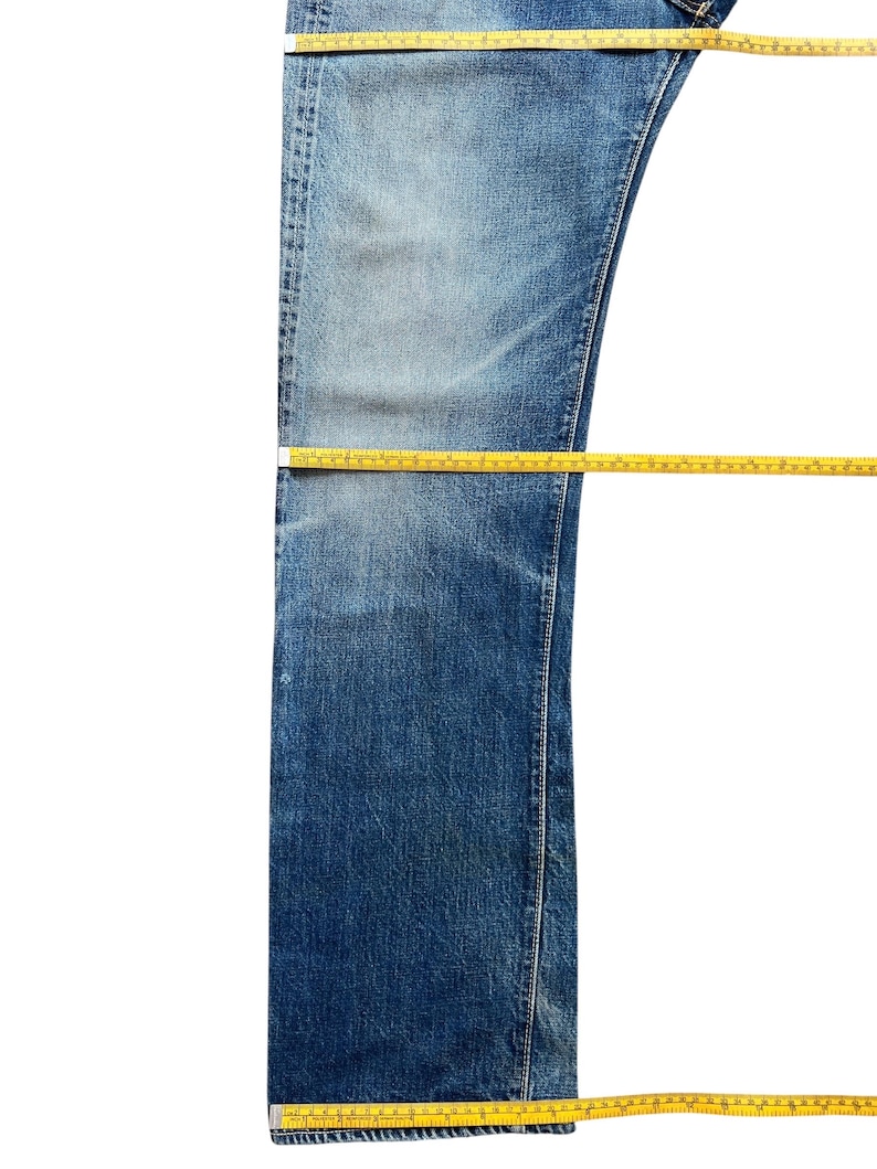 以下が含まれることがあります： 色あせたウォッシュ加工の青いデニムジーンズ。黄色のメジャーで測定されています。ジーンズは白い背景に平らに置かれ、片方の脚の前面が表示されています。メジャーはジーンズの長さと幅を示しています。