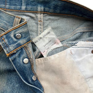 May include: Close-up of blue denim jeans with visible stitching and metal rivets. A tag with the text "W31 L32 501-0000" is visible. The jeans have a classic five-pocket design and a button closure.