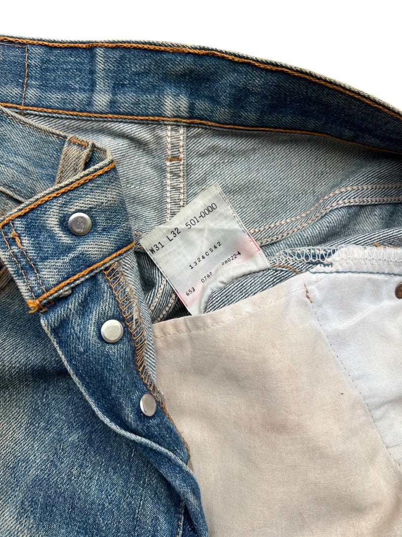 May include: Close-up of blue denim jeans with visible stitching and metal rivets. A tag with the text "W31 L32 501-0000" is visible. The jeans have a classic five-pocket design and a button closure.